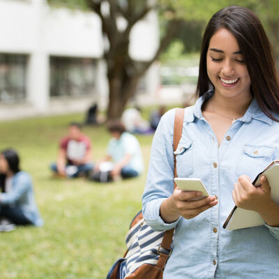 A female student walking outdoors holding her books and looking at her phone